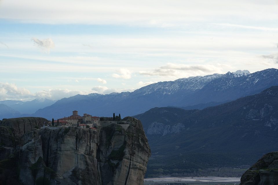 A monastery of tiled roofs perched on the flat top of a sheer rock tower, a valley and snow-streaked mountains behind in soft light