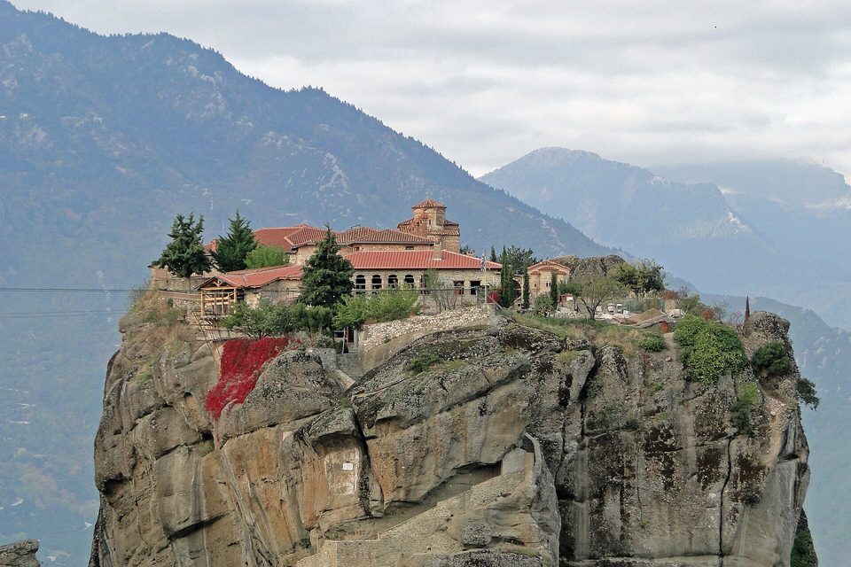 Monastery buildings and cypresses on top of a rounded, knobbly grey rock, red creeper on its face and steps cut into the rock below