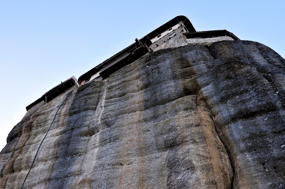 Looking up a smooth grey rock wall to monastery buildings built flush on its top edge against a pale blue sky, a cable running up the rock