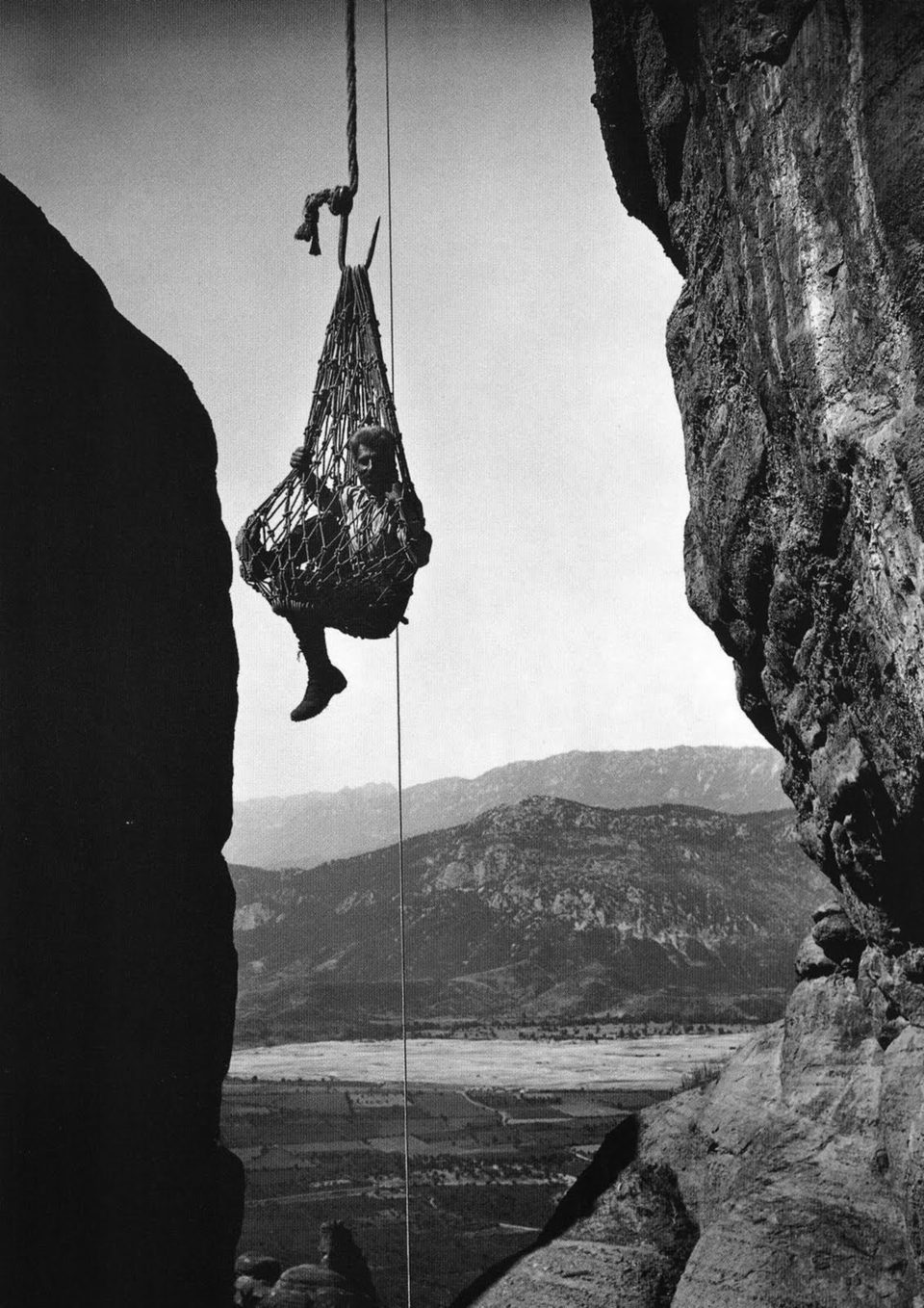 A black-and-white photograph of a man sitting in a rope net hanging from a hook between two cliffs, a valley and mountains far below