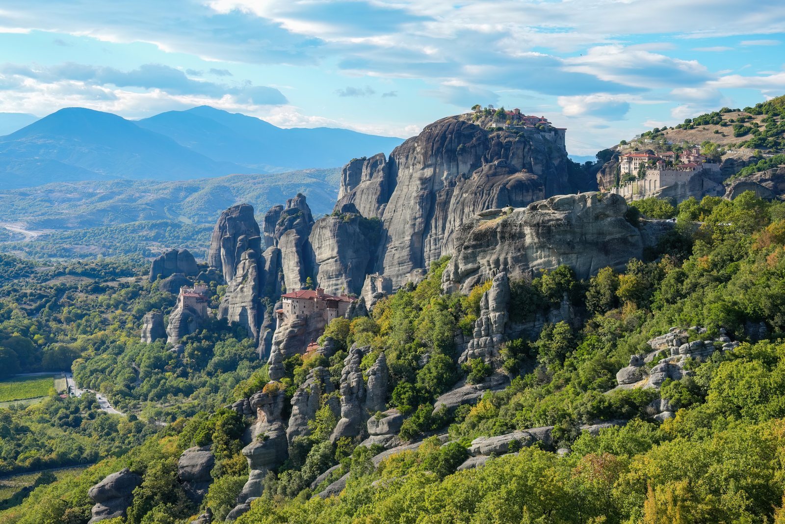 Tall grey conglomerate rock columns rising out of wooded slopes, monastery buildings standing on two of the summits, mountains beyond