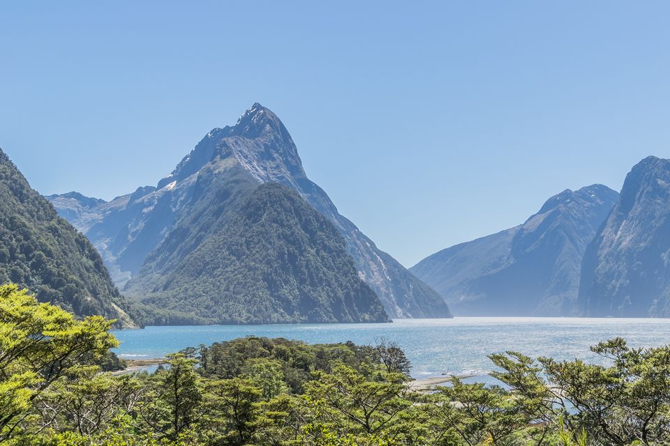A pointed mountain and a far shore across calm pale water, framed by green leaves at the left and right
