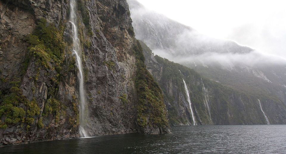A dark cliff running with white waterfalls above black water, more falls and mist on the far wall
