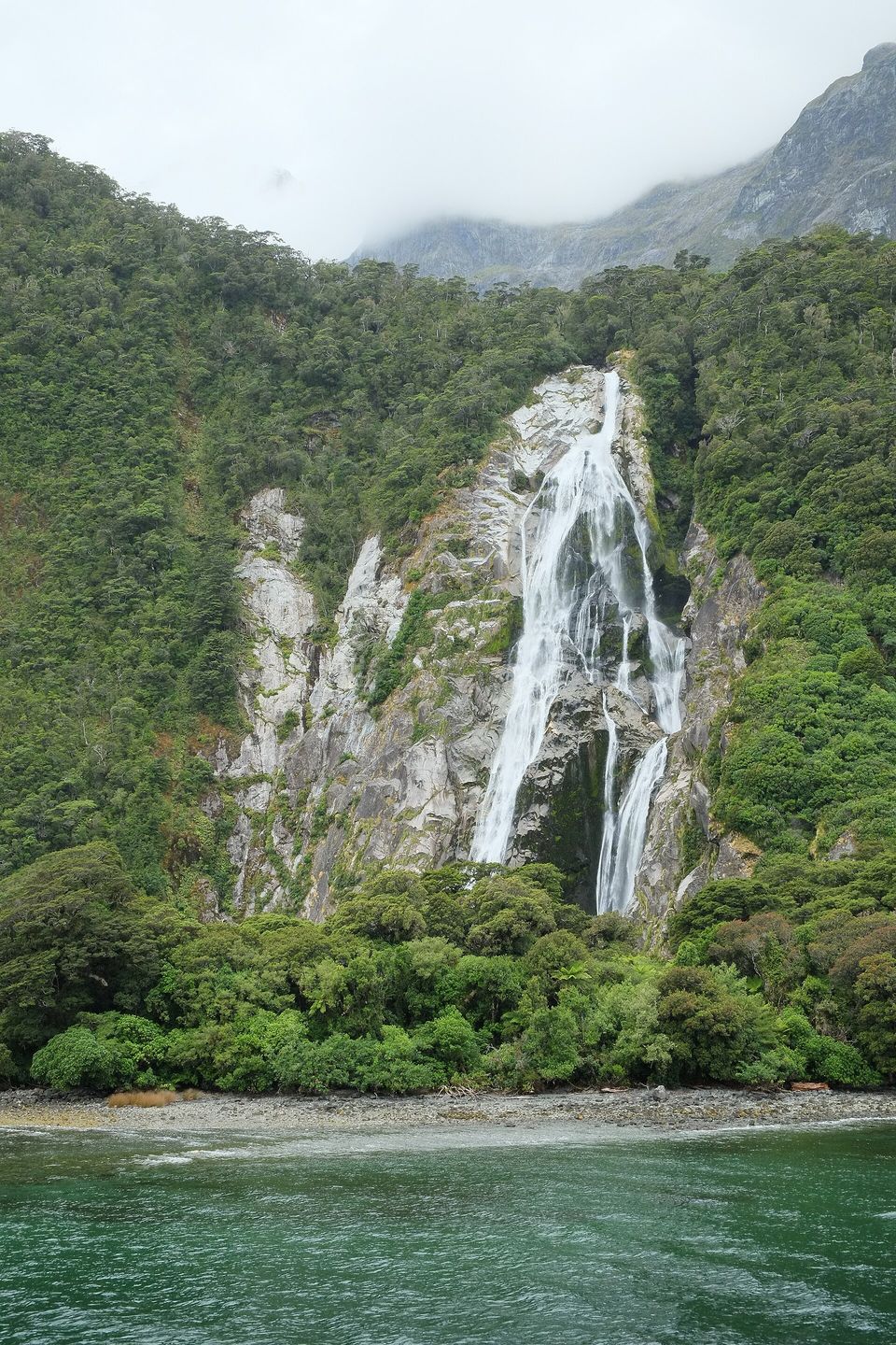 A thin white waterfall falling down a pale rock face streaked with dark, between green forest slopes above the water of the fiord