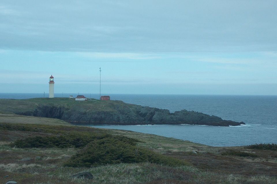 A white lighthouse with a red lantern and red-roofed buildings stand on a low green headland above a grey sea, heath and boulders in front