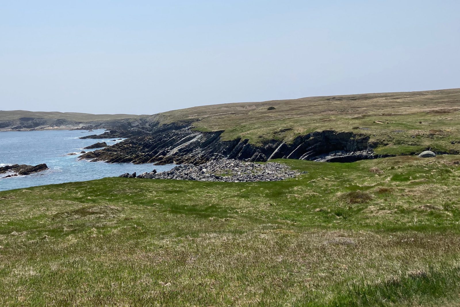 Green barrens fall to a low headland of steeply tilted dark rock beds and a shingle cove, the Atlantic beyond