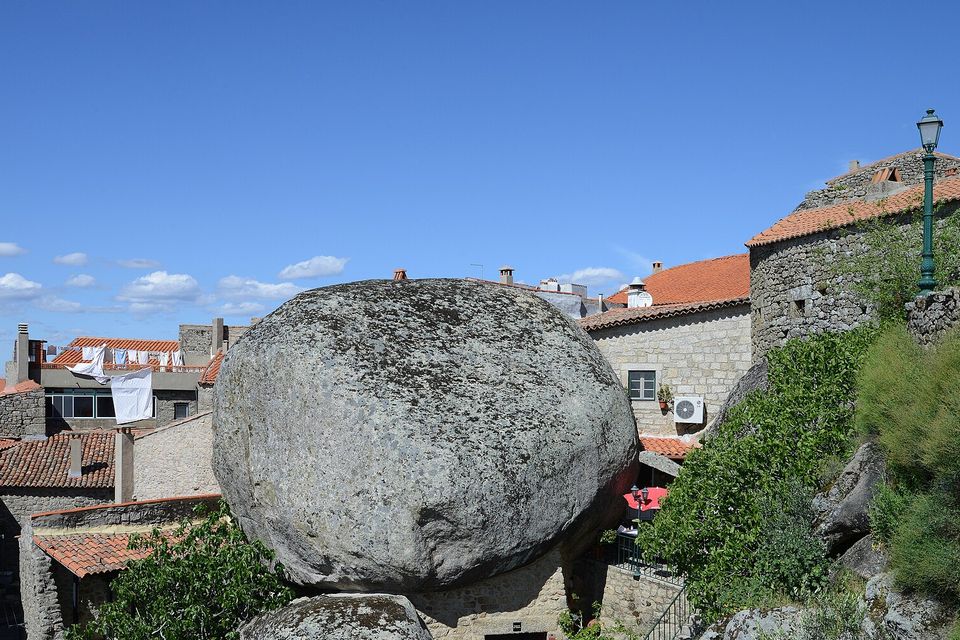 A vast rounded granite boulder rests on a smaller one, a stone house wedged beneath them, terracotta roofs and stone walls all around under a blue sky