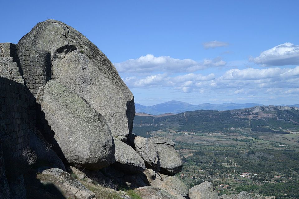 Huge granite boulders lean against a dry-stone castle wall on a cliff edge, with farmland, hills and distant mountains under white cloud beyond