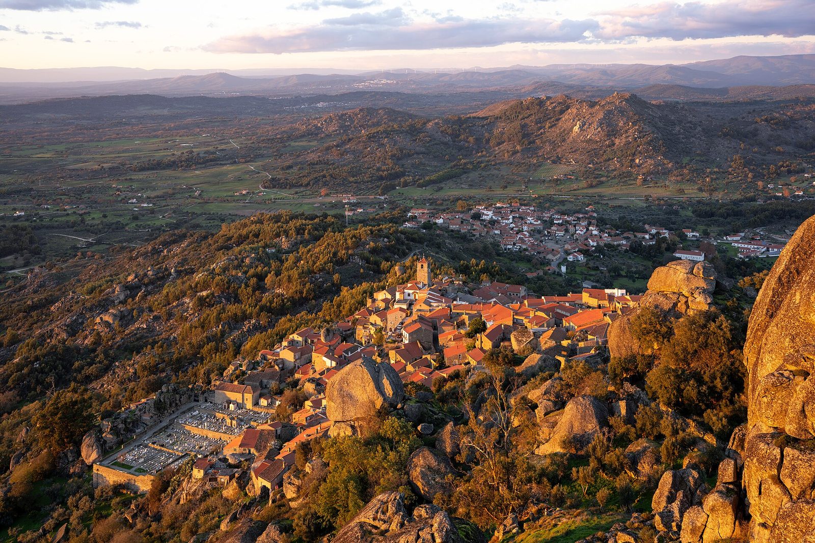 Golden-hour light from the castle rock: terracotta roofs and a clock tower tumble down a slope of giant granite boulders, the cemetery's white tombs at lower left, the plain of Beira Baixa beyond under a pink-and-blue sky