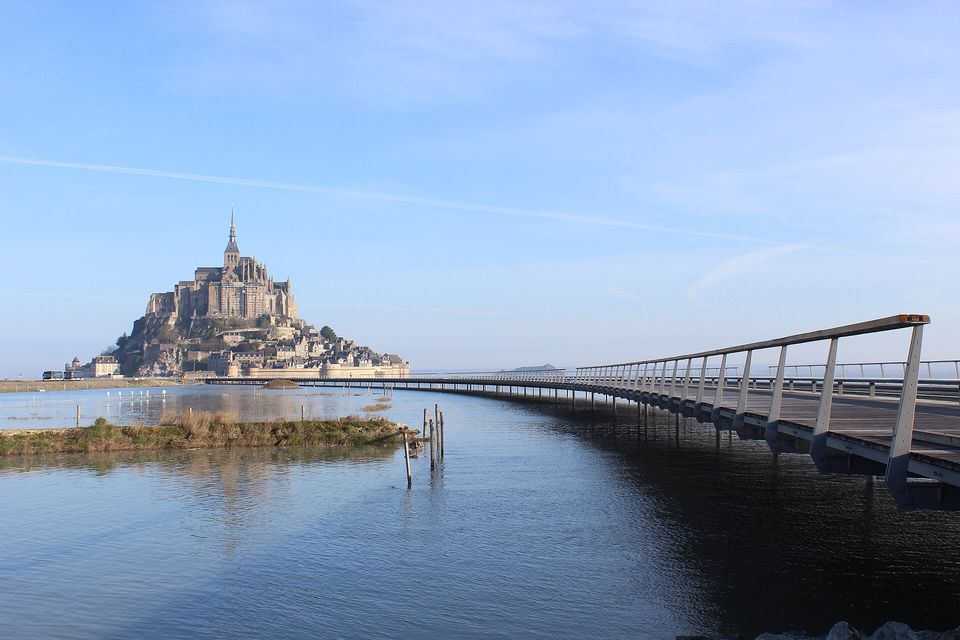 A rocky island with an abbey and village far off across calm blue water, a long raised walkway with a railing curving toward it from the right, pale sky above