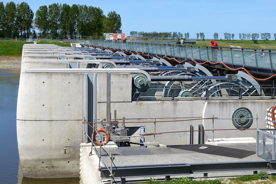 A pale concrete dam with steel gate wheels and railings above still water, a long glass-sided walkway running off to the right, trees and green meadow behind