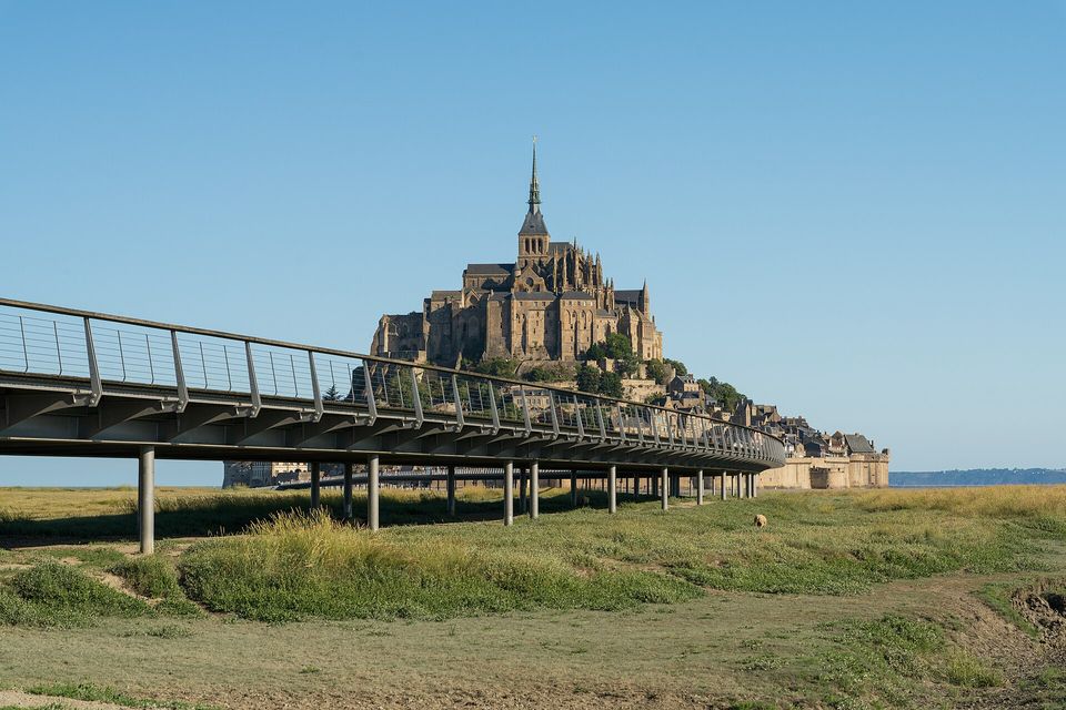 A slim raised bridge with a glass balustrade on thin round pillars crossing a grassy meadow toward an abbey-topped island, clear blue sky