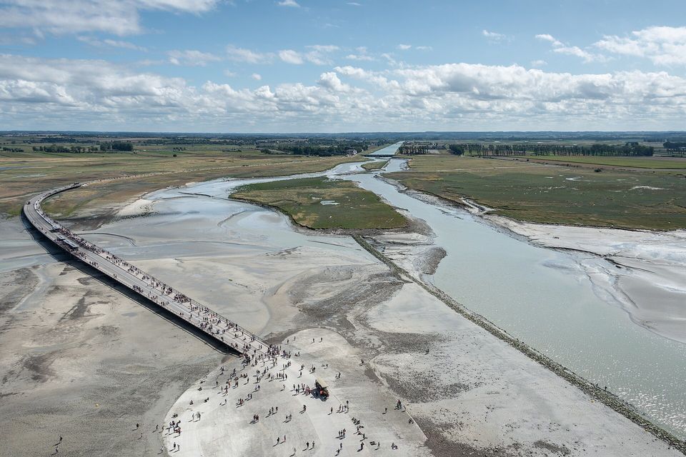 A view down onto a long straight walkway across pale tidal sand beside a channel of water, small groups of walkers on the sand at the near end, green marsh and a cloudy sky beyond
