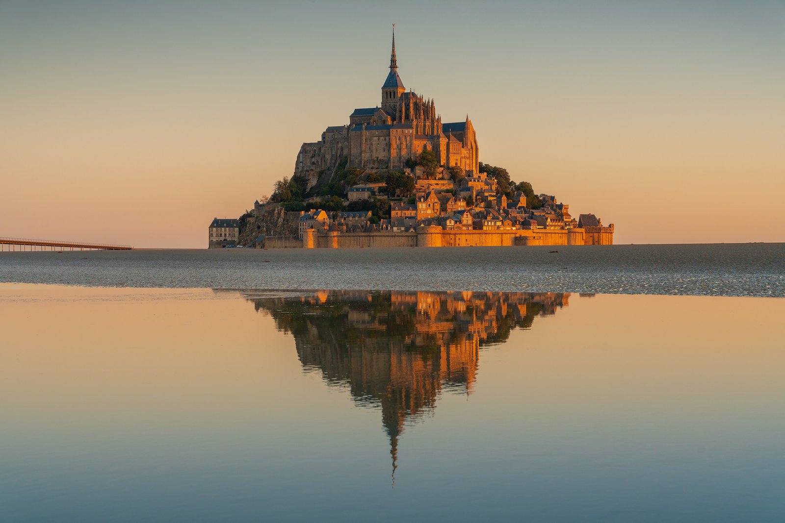 A rocky island crowned by an abbey with a tall spire and a huddle of houses below, lit gold at sunrise and mirrored in a sheet of shallow water, a slim raised bridge entering from the left edge