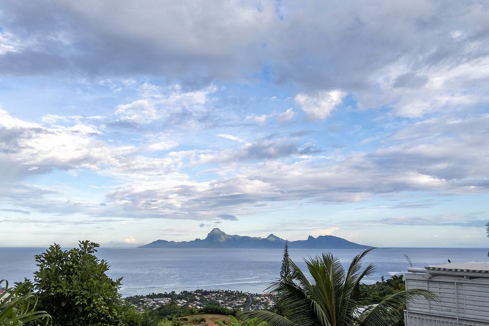 A distant jagged island on the sea horizon under a wide sky of broken cloud, with palms and a white balcony in the foreground and a town below