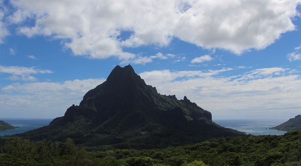 A dark mountain spire silhouetted against bright cloud, with a bay showing at each side and forest in front