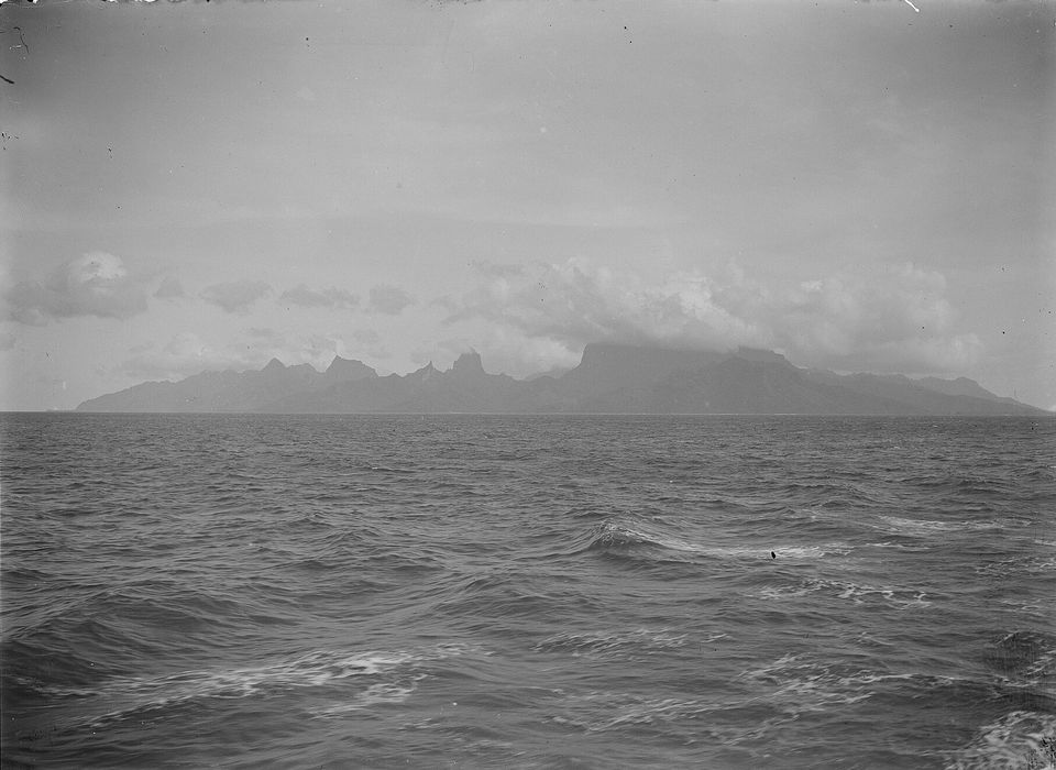 A black and white view across a rough sea to a low, jagged island under cloud