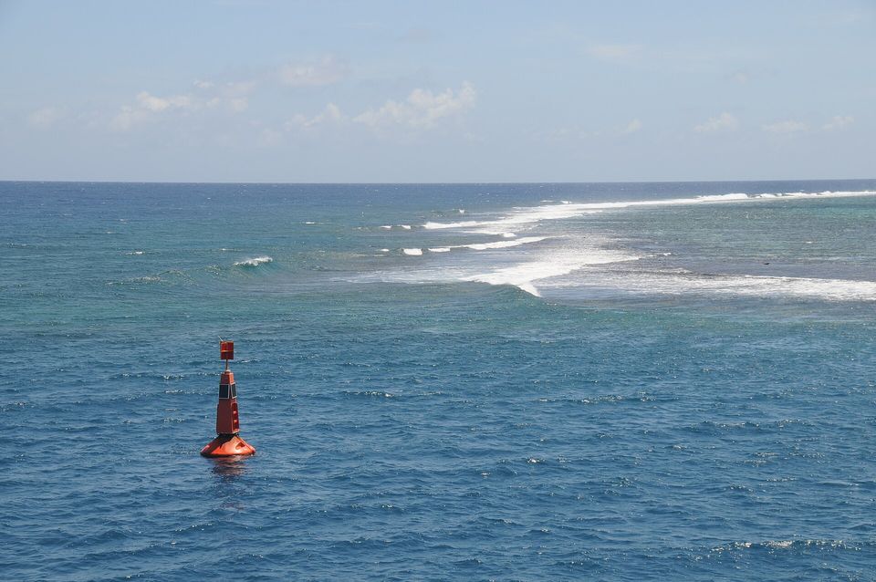 A red channel buoy floating in deep blue water, with white surf breaking along a reef beyond it under a pale sky