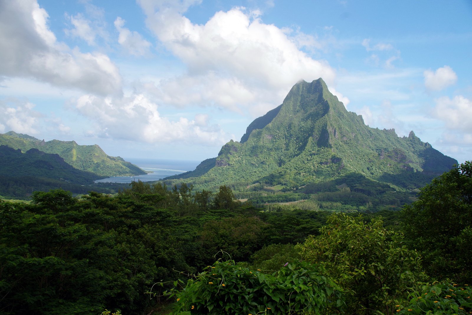A steep green mountain spire rising above dense forest, with a sliver of sea between two ridges at the left, under bright broken cloud