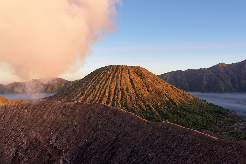 A ridged green and orange cone lit by low sun beyond a dark crater rim, a pink cloud of steam at the left and mist lying in the caldera behind