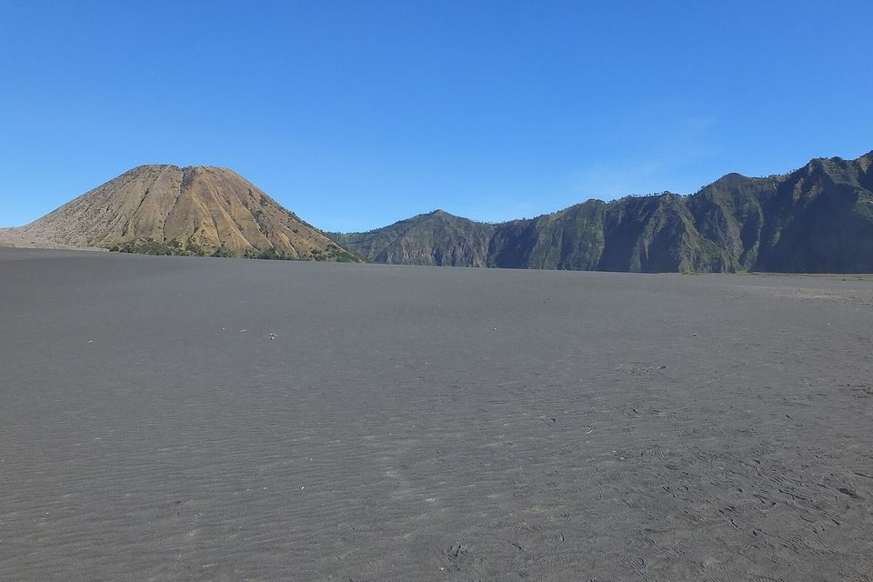 A wide plain of dark grey wind-rippled sand under a deep blue sky, a bare golden-brown cone at the left and a green ridged caldera wall running off to the right