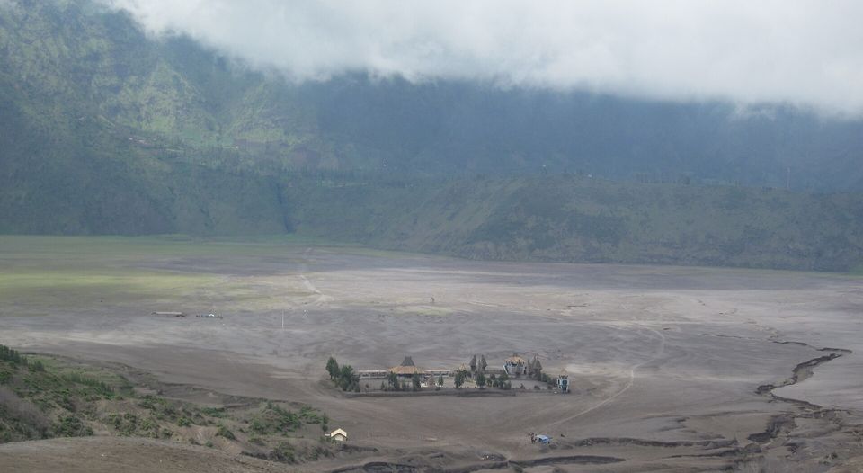 A small walled temple compound on a wide grey ash plain beneath a green caldera wall and low cloud