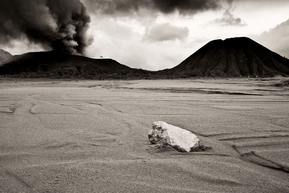 A black and white view across pale ash-rippled sand to two dark volcanoes, black ash rising from the left one under a heavy sky, a single pale boulder in the foreground