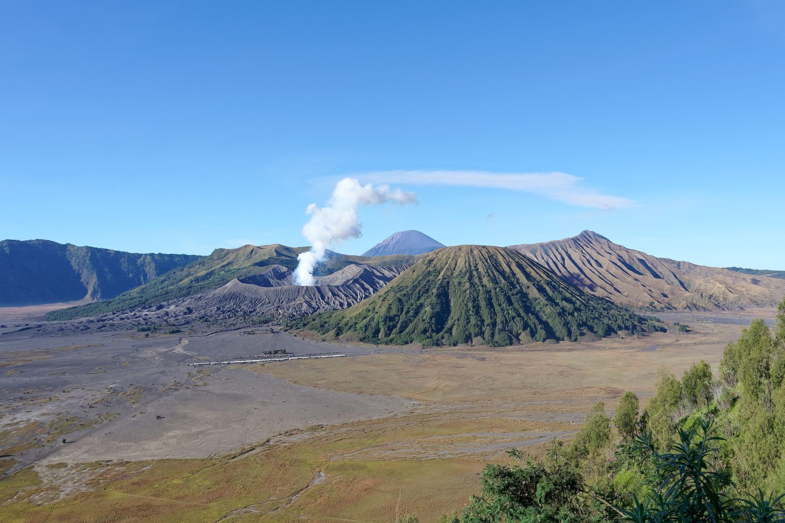 A plume of white steam rising from a grey volcanic crater beside a green ridged cone, a distant blue peak behind and a flat grey ash plain in front, under a clear blue sky