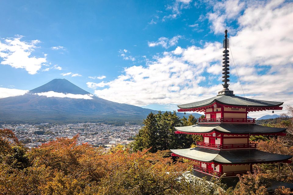 A red five-storey pagoda among autumn trees on a hillside, a town below and a distant snowless Mount Fuji under blue sky