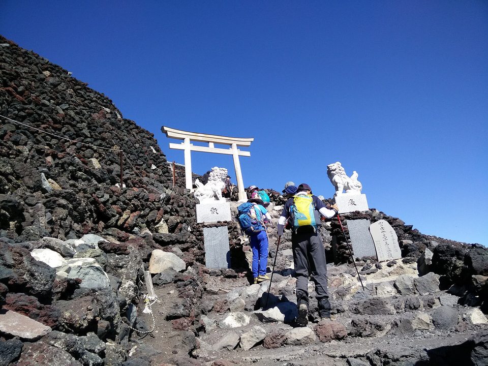A small white torii gate on dark volcanic rock above two hikers seen from behind, under a deep blue sky