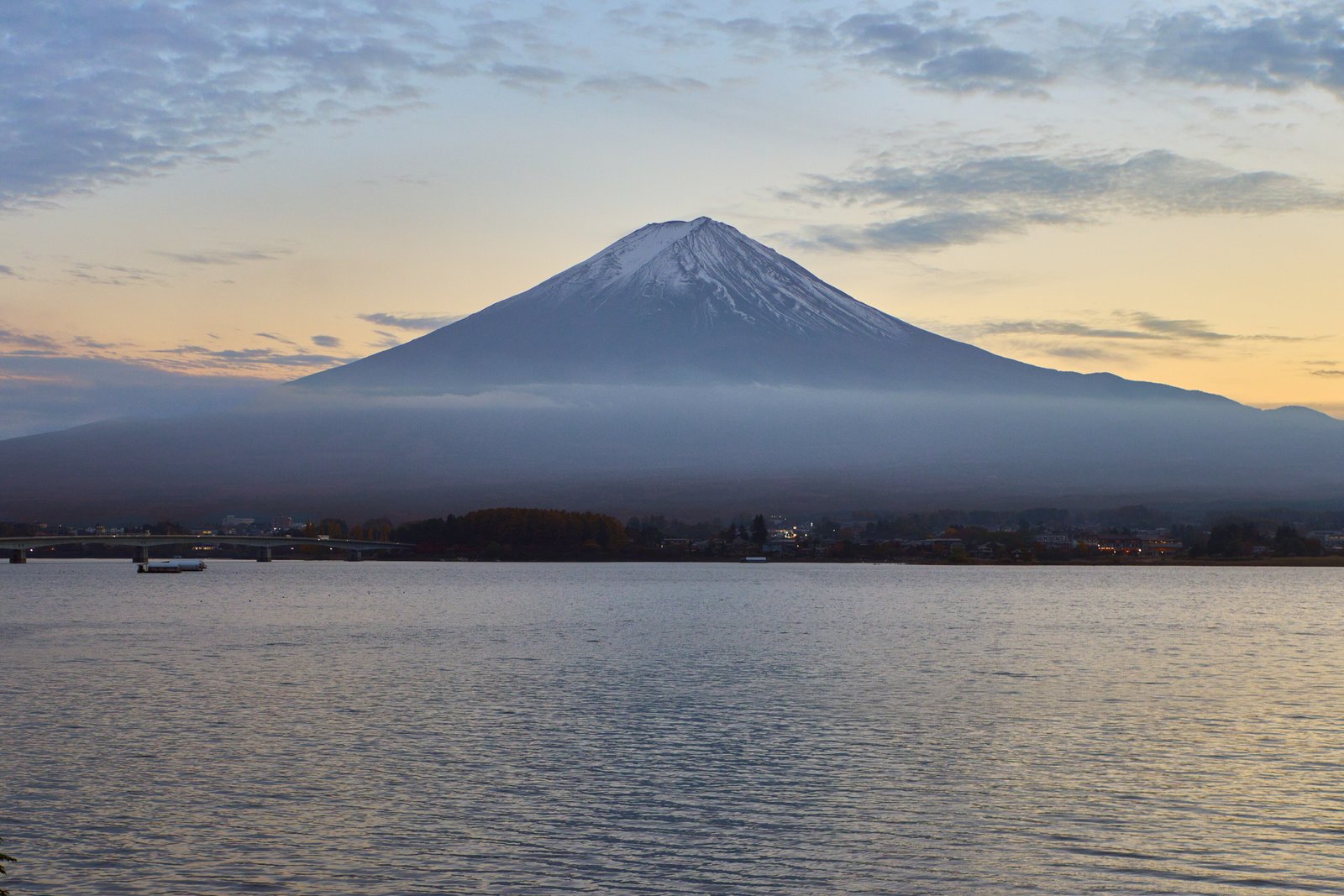 A snow-capped cone-shaped mountain above a band of low cloud and a calm lake, in soft dawn colours