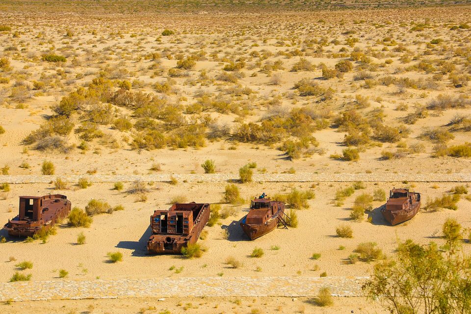Four rusted hulls standing in a row across pale sand and scrub, a paved path along the foreground, seen from a height
