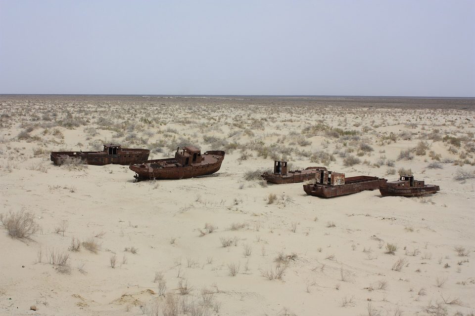 Five rust-brown boats scattered across a flat pale sand plain with low scrub, a hazy horizon far behind