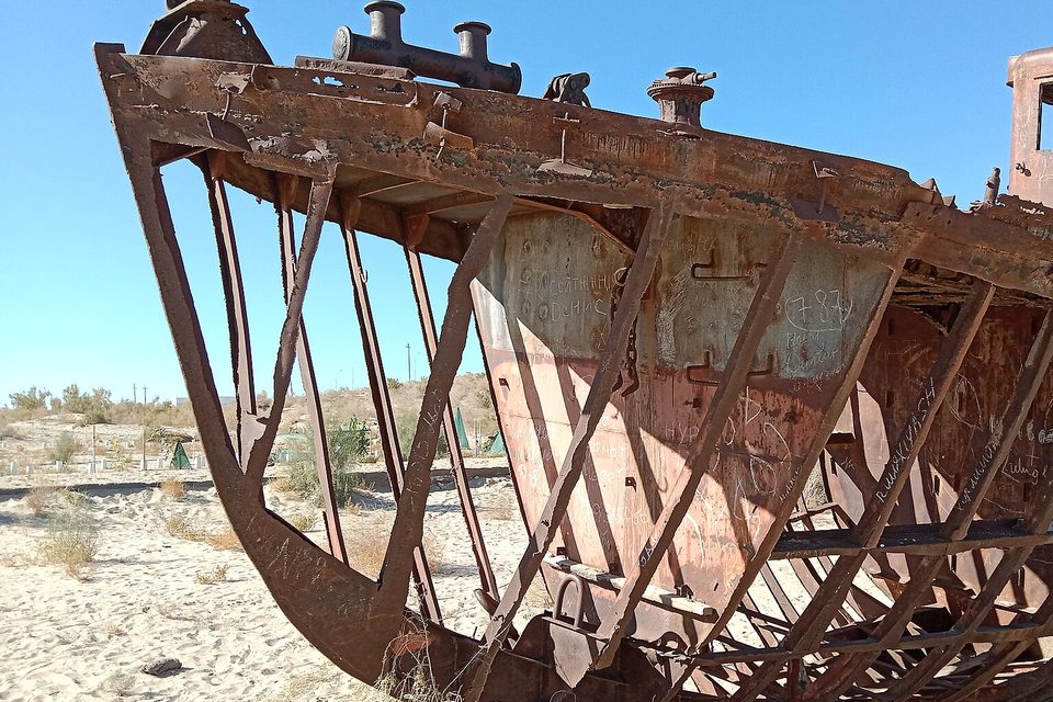 A rusted ship hull seen from the bow, its plating gone to leave curved ribs against a blue sky, sand and low scrub below