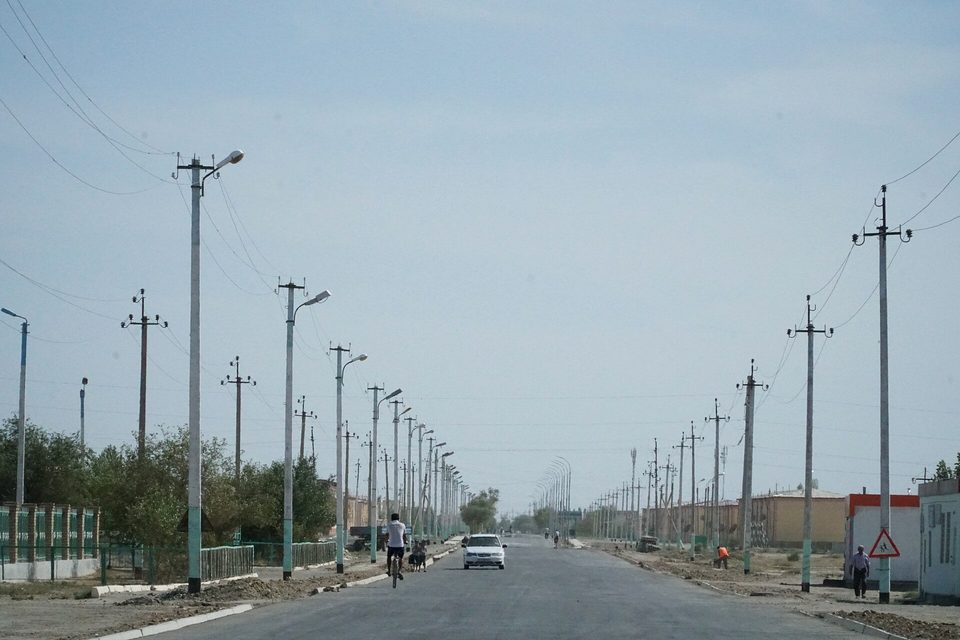 A wide empty road lined with lamp posts and power poles running to a hazy distance, a cyclist and a car on the road