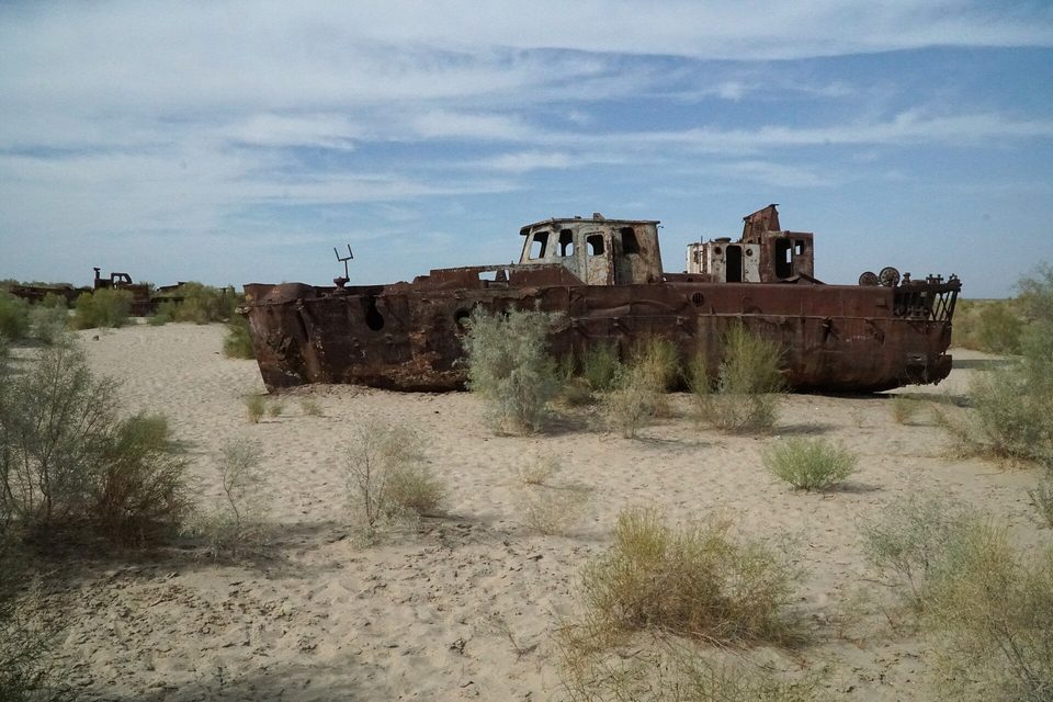 A long rusted hull standing broadside on pale sand among green desert shrubs, under a blue sky streaked with cloud