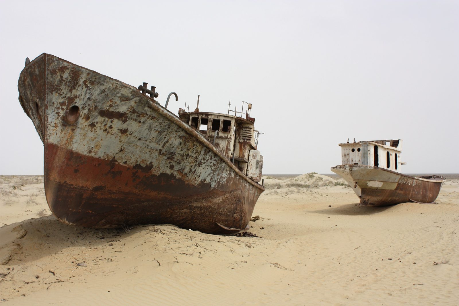 A rust-streaked trawler's bow resting upright in rippled sand, a second white-cabined hull beyond it, under a pale sky