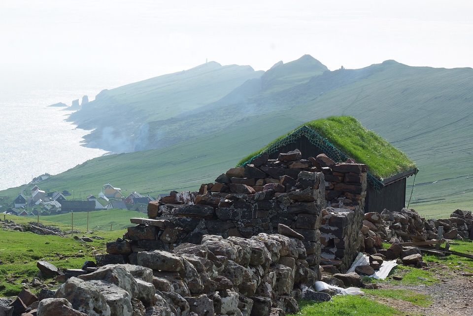 A turf-roofed stone building in the foreground, the village and green headlands of Mykines beyond