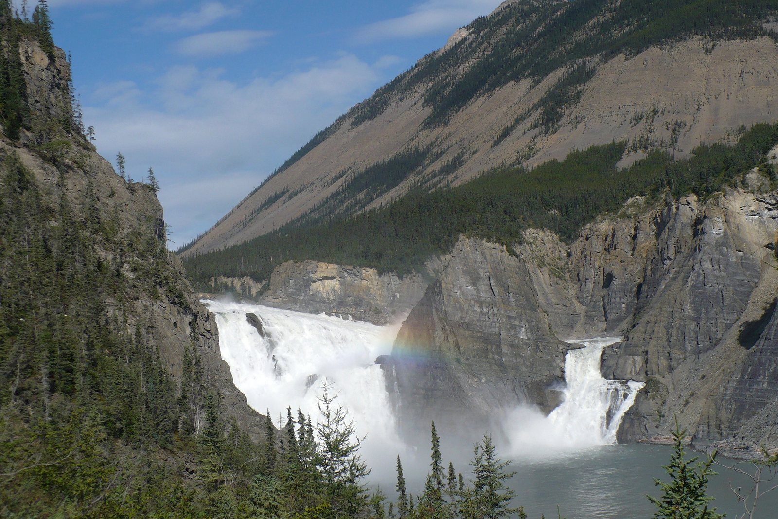 A river falls dropping in two sheets either side of a rock spire, a faint rainbow in the spray, forested canyon walls behind