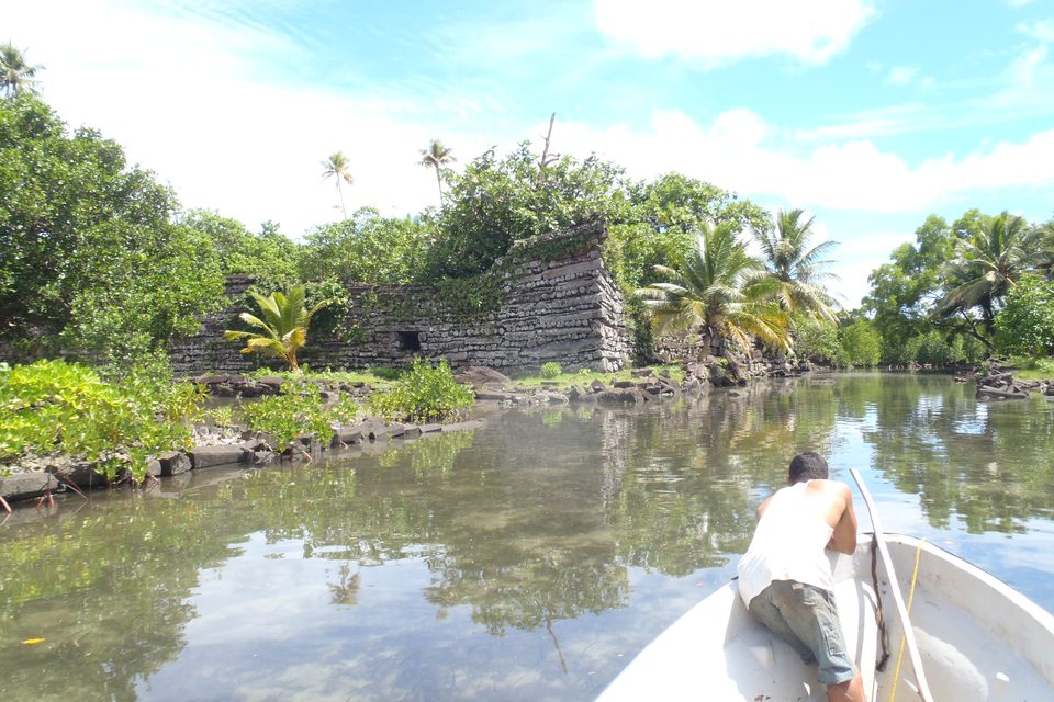 The bow of a small boat moving along a calm canal toward basalt walls half hidden by palms and bushes