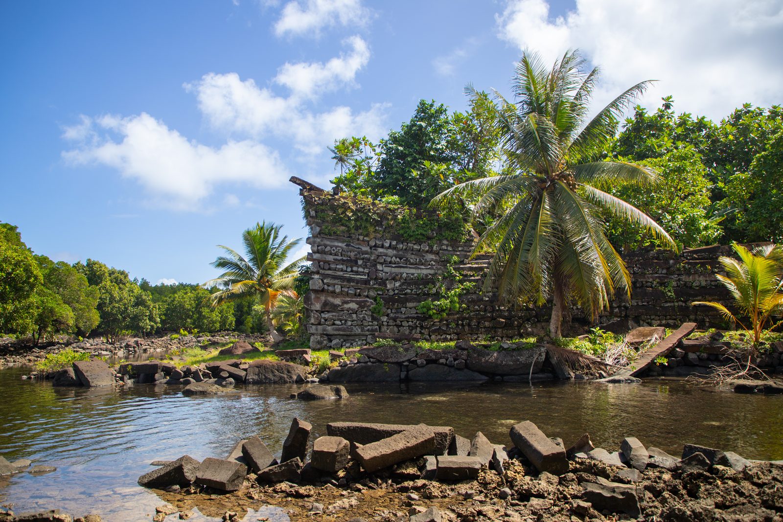 A wall of dark basalt columns stacked crosswise in courses, its corner sweeping upward, standing over a still tidal canal in low afternoon light, palms and blue sky above