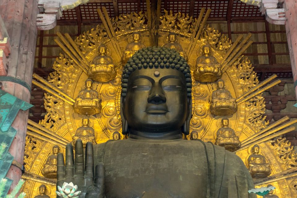 A bronze Buddha's face and raised hand in front of a gilded halo of small seated Buddhas, between red pillars