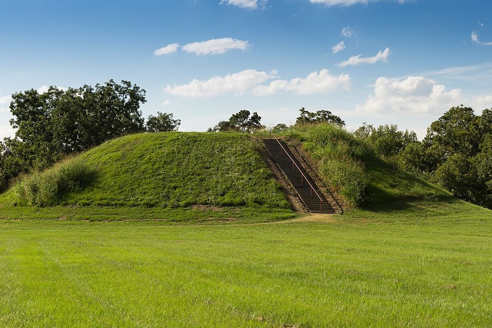 A broad flat-topped grassy mound with a steep stair up one side, trees behind and mown grass in front under a blue sky