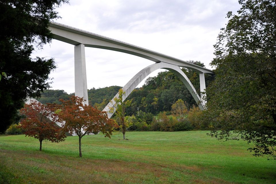 A long white concrete bridge on two shallow arches crossing a wooded hollow, red-leafed trees and a mown field below