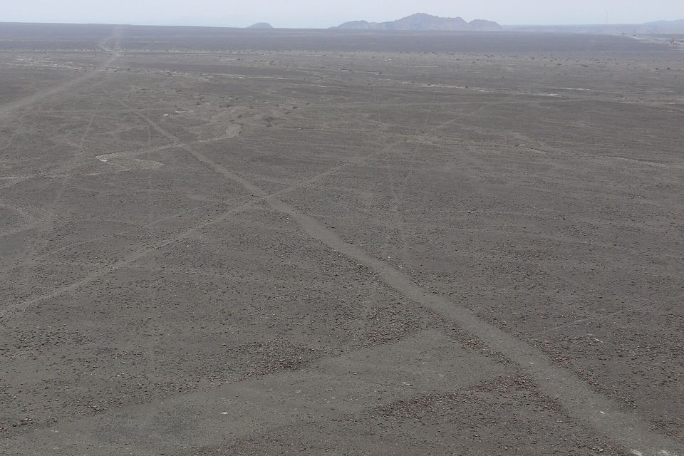 A flat grey stony plain with faint straight lines crossing it, seen from a small hill, low hills on the horizon