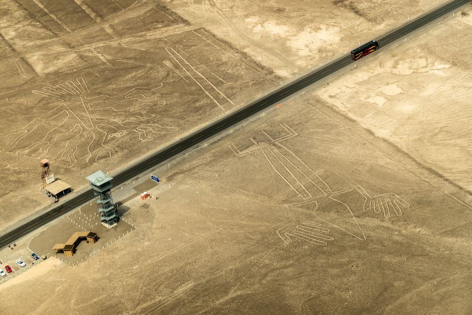 An aerial view of a straight highway crossing the stony pampa beside a roadside viewing tower, with the Tree and the Hands geoglyphs drawn on the ground