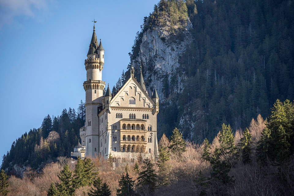 A pale stone castle with a tall round tower and a gabled front on a wooded slope, bare trees in the foreground and a dark forested cliff behind, under a violet-blue sky