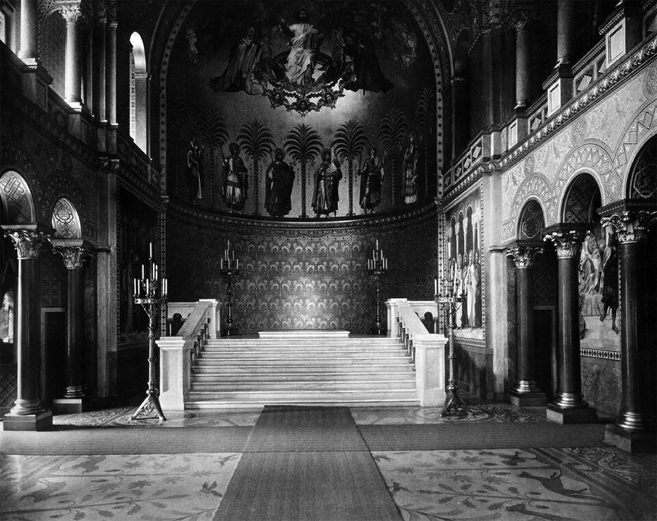 A black-and-white photograph of a tall hall ending in an apse painted with figures and palm trees, a flight of marble steps rising to an empty platform between two tall candlesticks