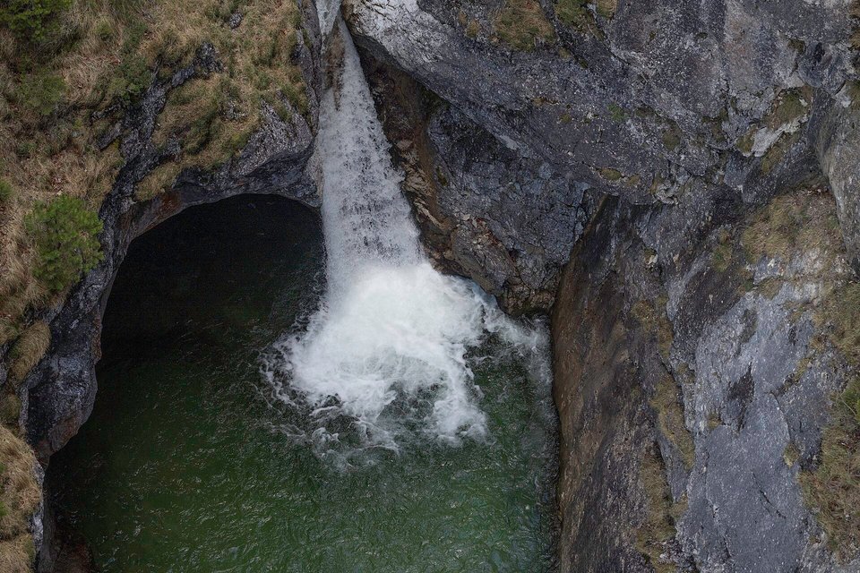 A white waterfall dropping into a dark green pool inside a narrow rock gorge, seen from above