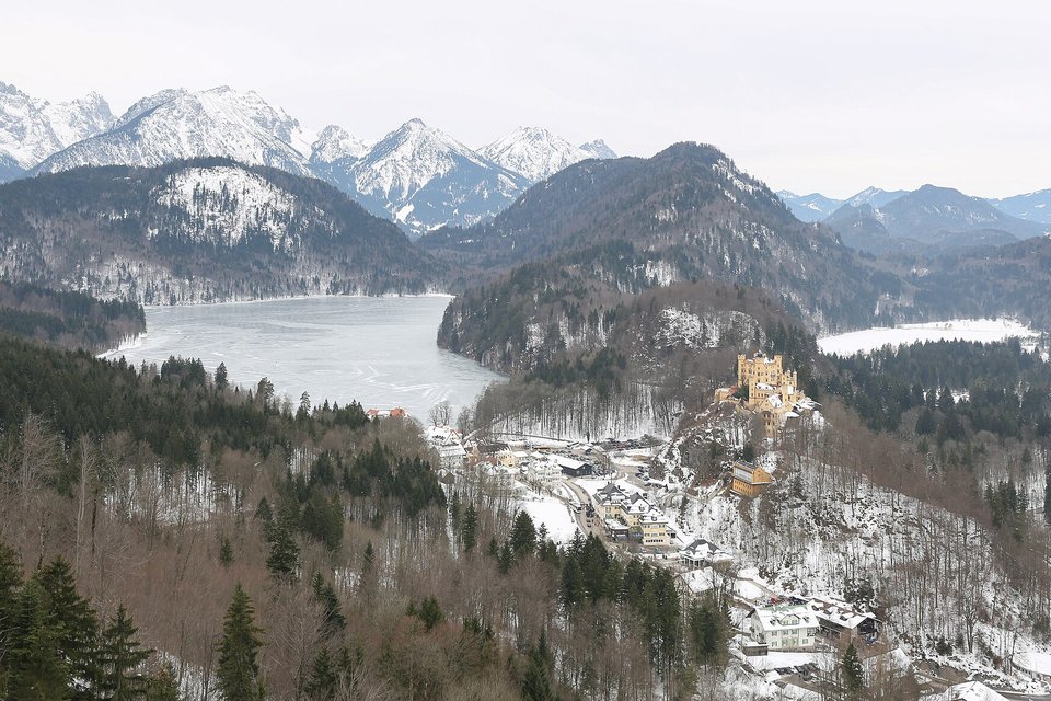 Snow-dusted forested ridges and a frozen lake, a small yellow castle on a wooded hill beside a village, pale mountains in the distance